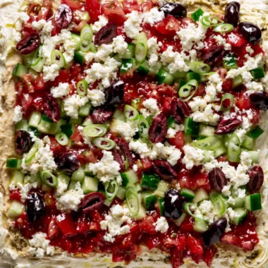 Greek-inspired layered dip with Mediterranean ingredients in a colorful presentation.